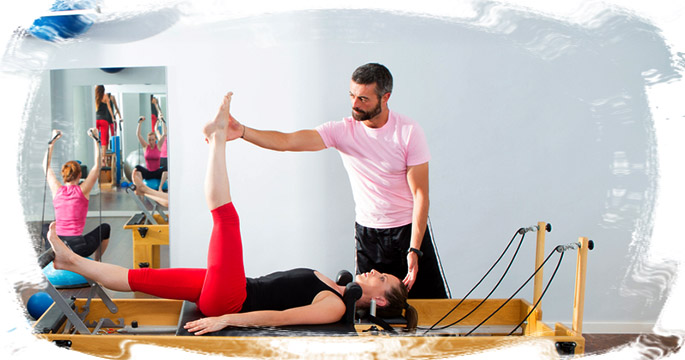 Monitor ayudando a una chica a realizar pilates máquina y otras chicas realizando pilates suelo